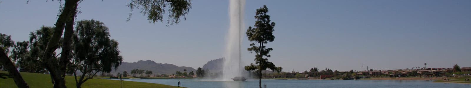 A water fountain in the center of a lake with trees, grass and clouds