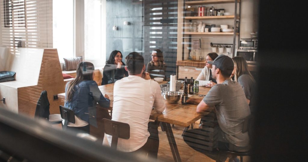 A group of seven young adults sit around a casual kitchen table.