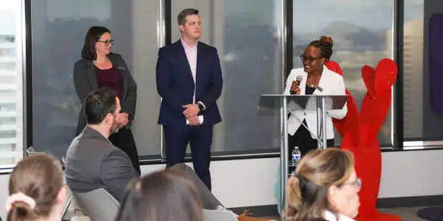 Two women and a man stand in front of an audience speaking into a microphone.
