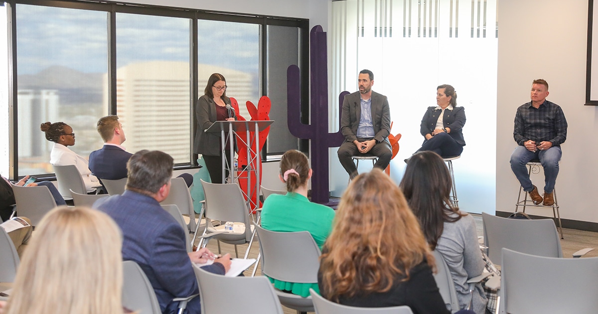 Three people sit in front of an audience with a moderator to the left.