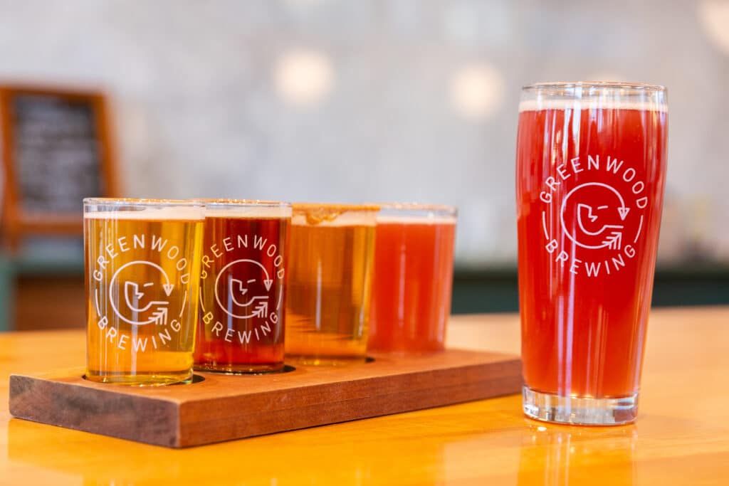 Four smaller beers displayed on a wooden plate for a flight sit on a bar with one larger red-pink colored beer.