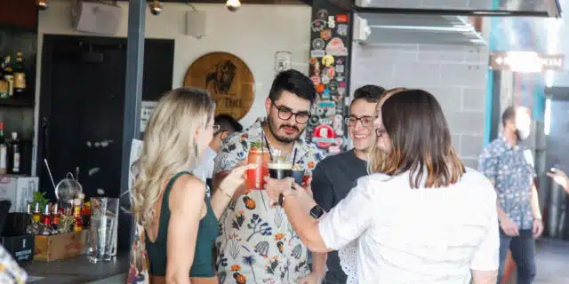 Four people facing each other and standing in front of an outdoor bar cheers with their cocktails.