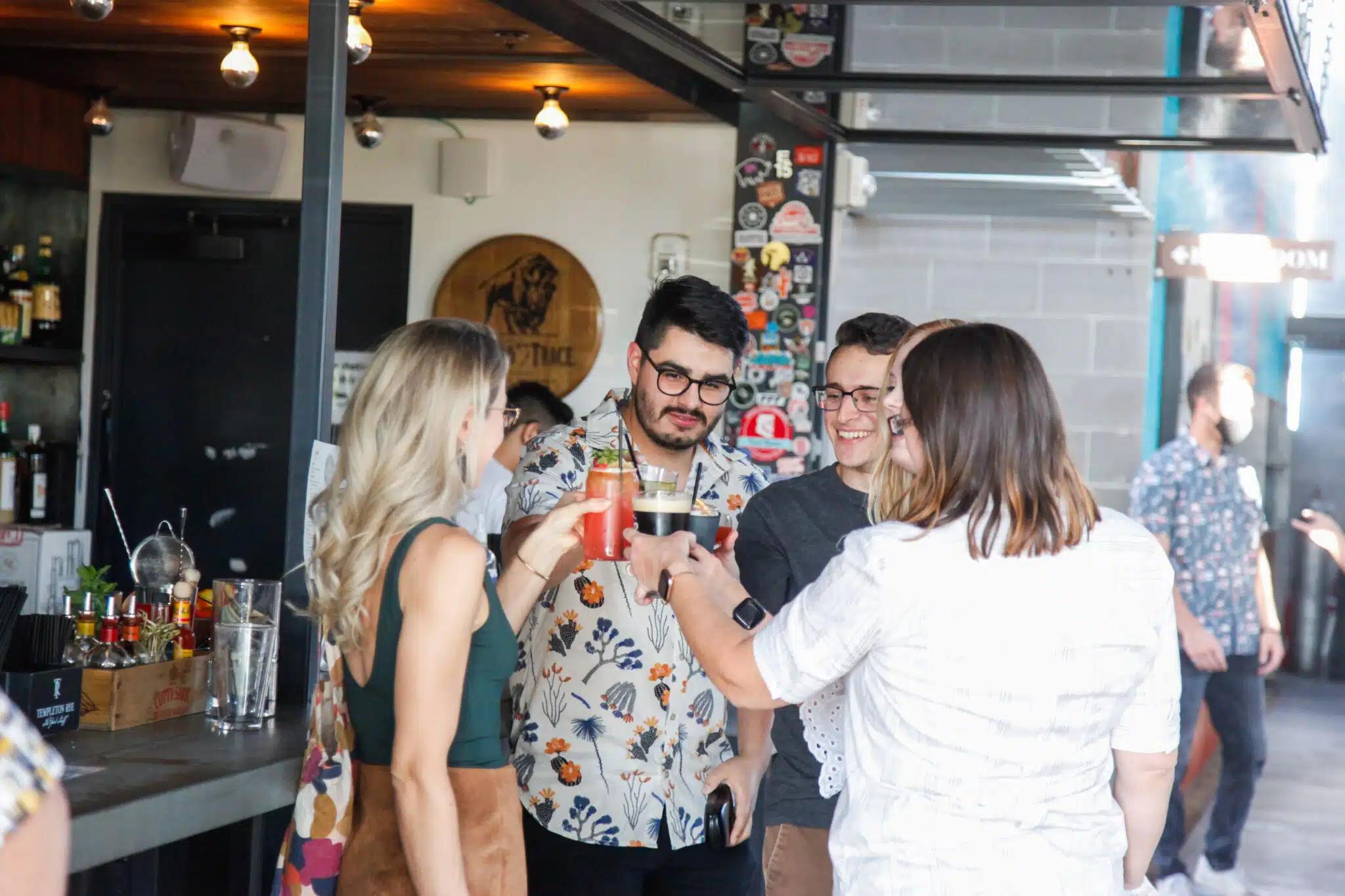 Four people facing each other and standing in front of an outdoor bar cheers with their cocktails.