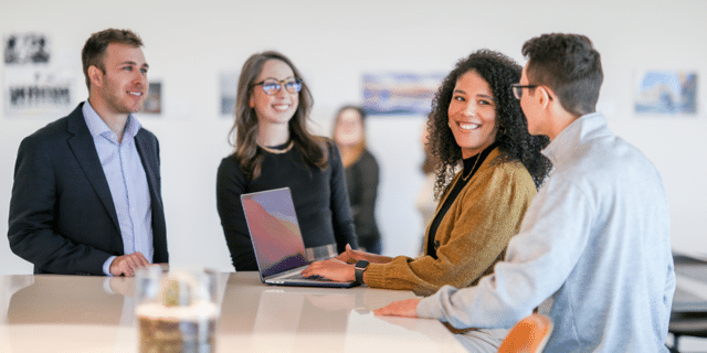 Four people are smiling and standing at a high table looking at a laptop.