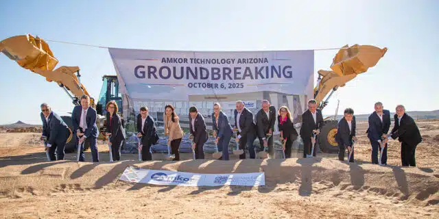 A line of people gather dirt onto a shovel in front of an Amkor sign and two construction vehicles.
