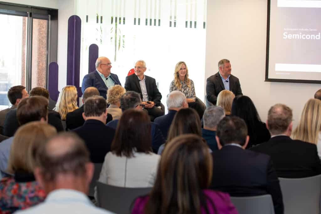 A crowd of seated people in the foreground listens to speakers on high chairs in the background.