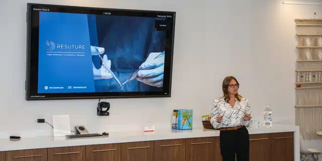A women speaks in front of a wall-mounted television displaying a slide from Resuture