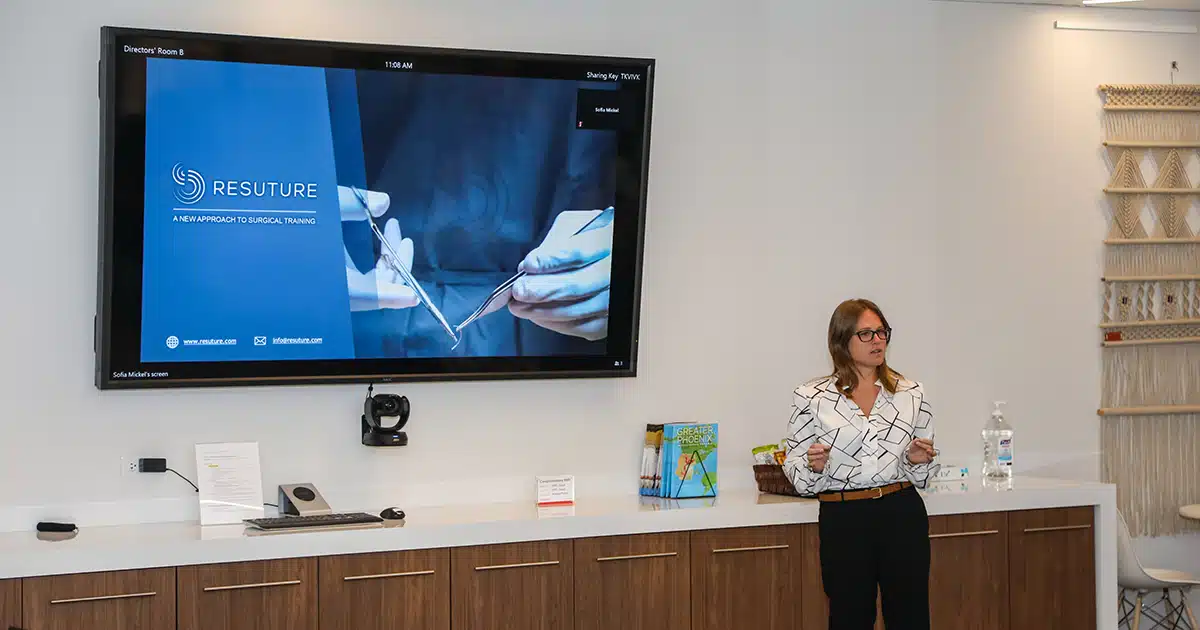 A women speaks in front of a wall-mounted television displaying a slide from Resuture