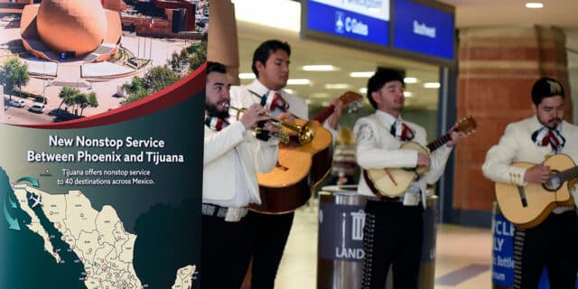On the left third, a tall sign with a picture of Tijuana and outline of the Mexico map announce the new American Airlines route between TIJ and Phoenix. To the right, four mariachi musicians play instruments.