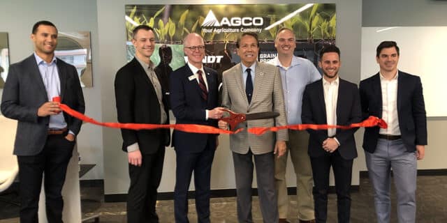 Seven men stand behind a red ribbon with an oversized pair of scissors. An AGCO sign is directly behind them on an office wall.