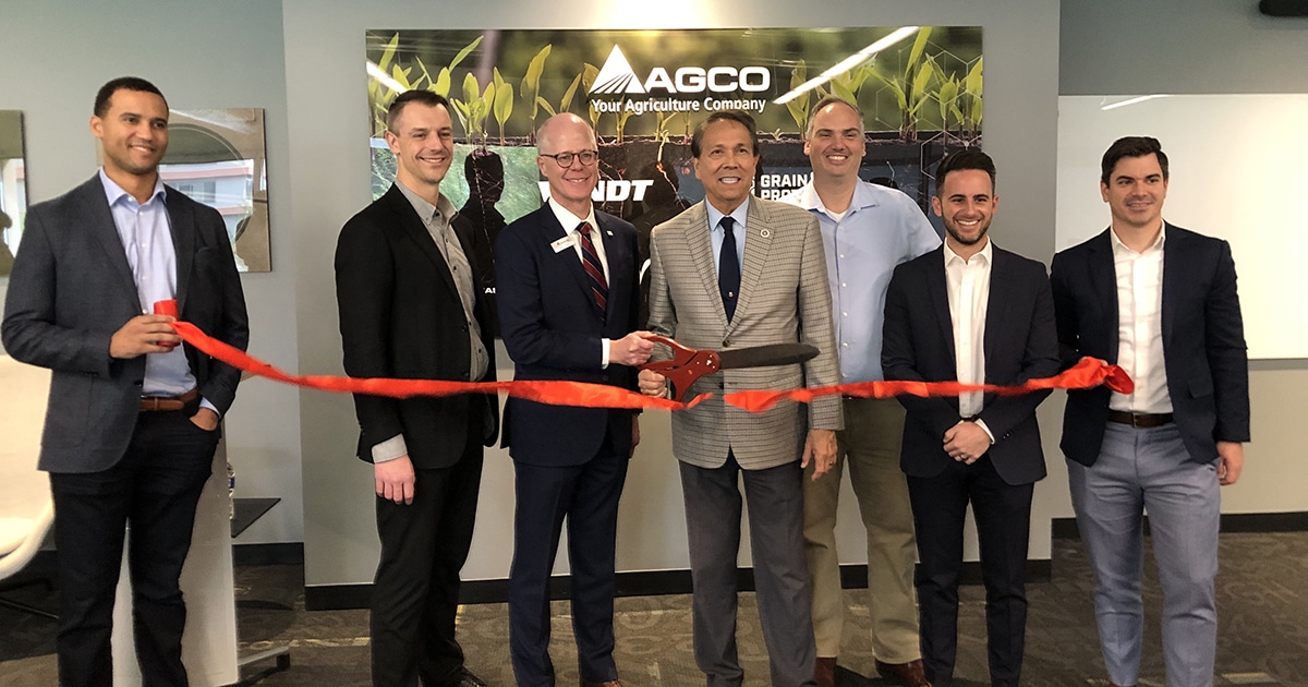 Seven men stand behind a red ribbon with an oversized pair of scissors. An AGCO sign is directly behind them on an office wall.