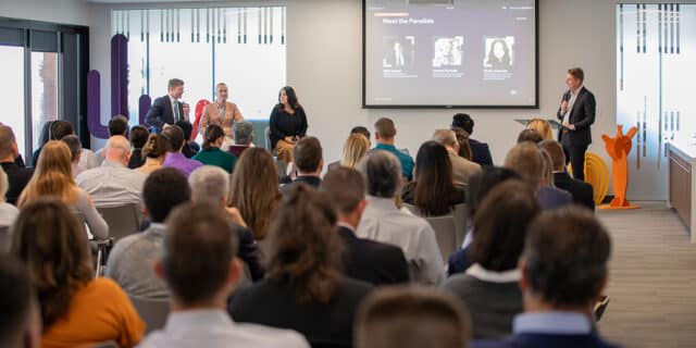 In the foreground, a group of seated individuals listen to a panel of three people seated the left and a man standing on the right.