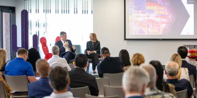 In the foreground, a group of people listen to a seated man and woman speak.