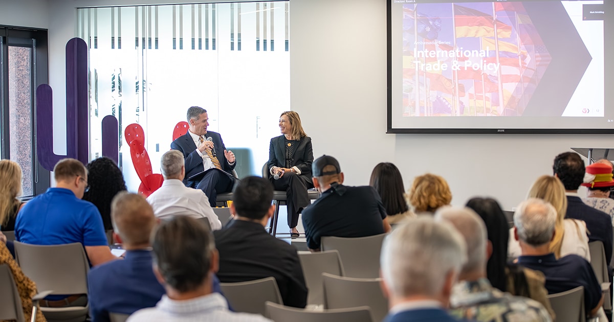 In the foreground, a group of people listen to a seated man and woman speak.