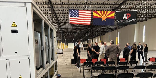 In the background, people talk under three flags: USA, Arizona and one with the Apex Power logo. On the left in the foreground is a completed converter manufactured by the company.