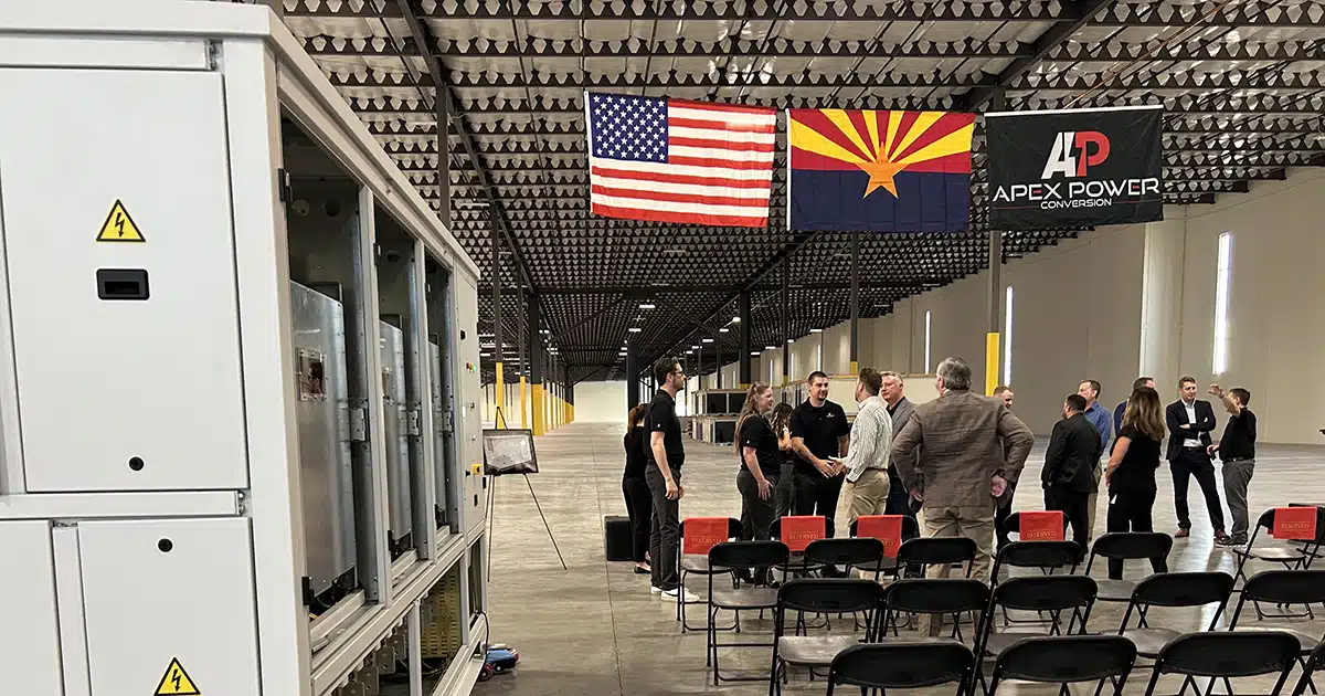 In the background, people talk under three flags: USA, Arizona and one with the Apex Power logo. On the left in the foreground is a completed converter manufactured by the company.