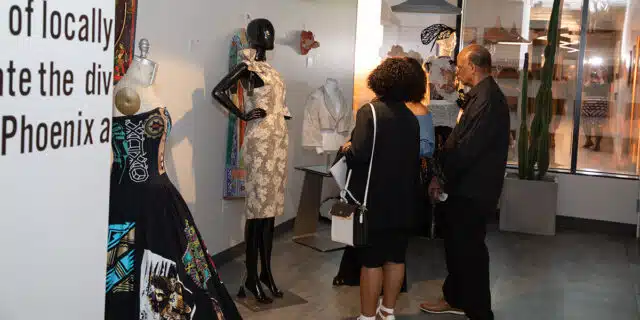Three people standing close together look at a mannequin wearing a dress designed by an artist in GPEC's Color, Cloth & Culture art exhibit.