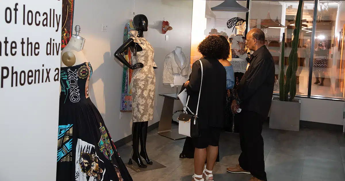 Three people standing close together look at a mannequin wearing a dress designed by an artist in GPEC's Color, Cloth & Culture art exhibit.