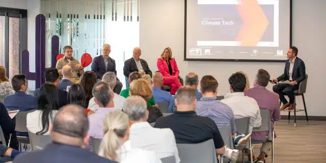 In the foreground, a group of seated people listen to a panel of speakers on high chairs in the background.