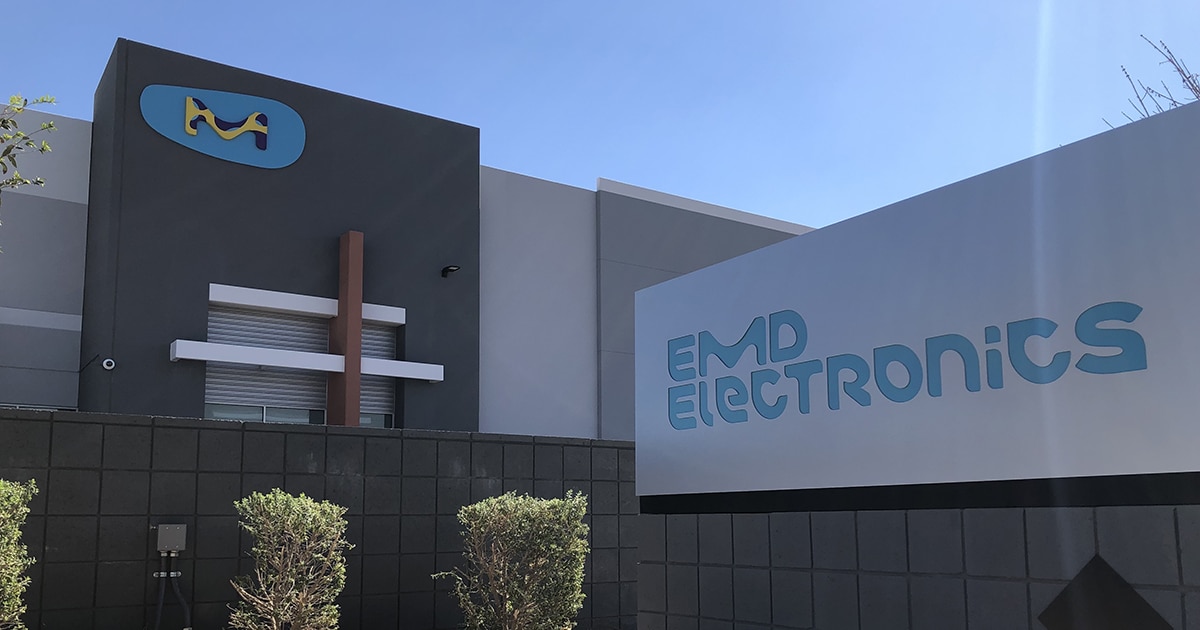The EMD building with a light blue logo in the background, and a sign with the company's full logo in the foreground.