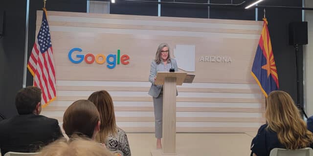 Gov. Hobbs speaks at a podium with the USA flag and google logo to the left and Arizona state outline and flag to the right.