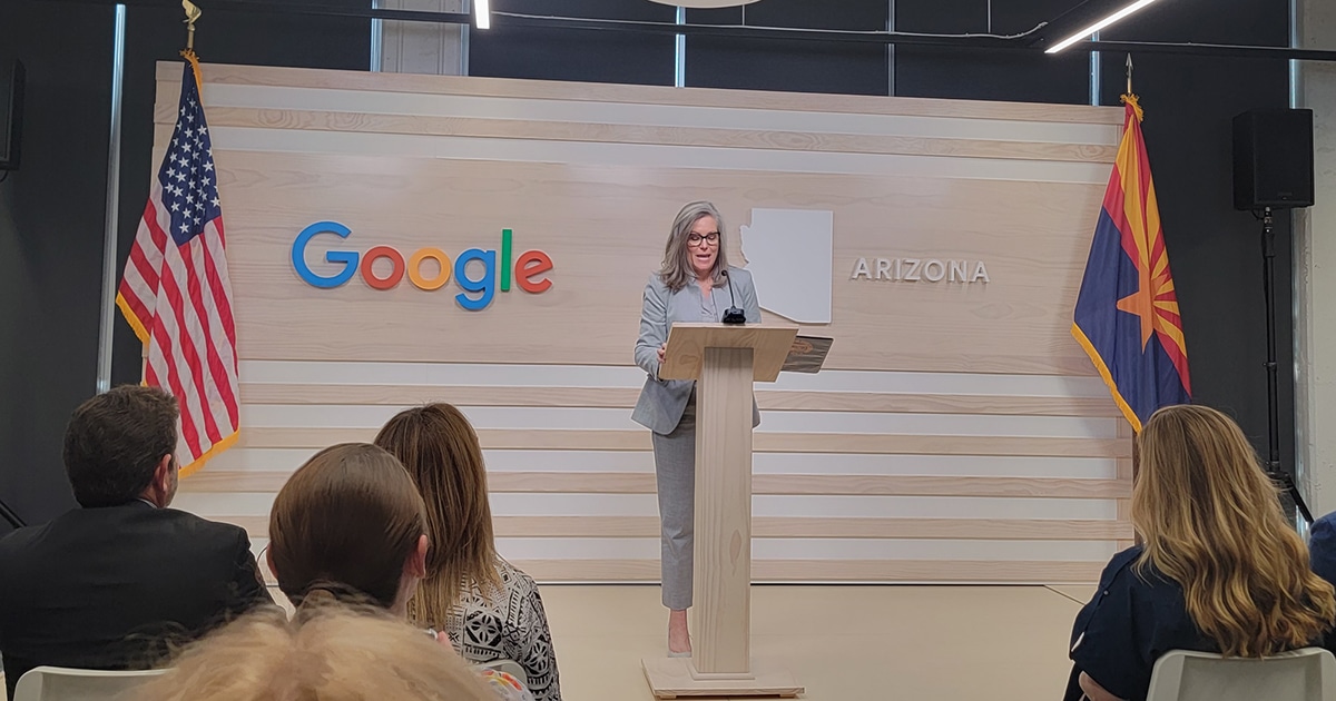 Gov. Hobbs speaks at a podium with the USA flag and google logo to the left and Arizona state outline and flag to the right.