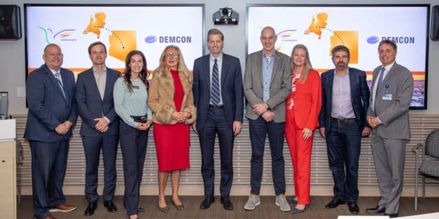 A line of well-dressed men and women pose for a photo in front of projector screens displaying the Hummingbird and DEMCON logos and the outlines of Holland and Arizona.