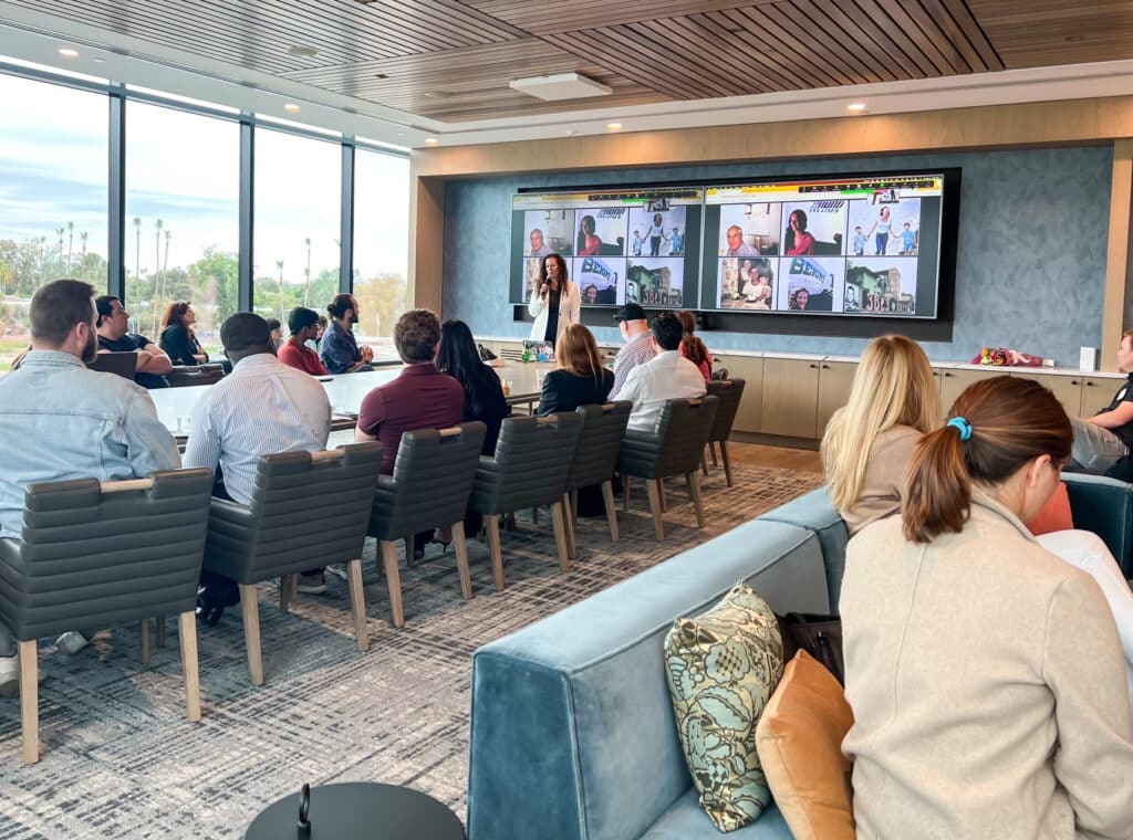 A group of people sit at a long table while, on the right side, a few more are on couches. They listed to a speaker at the front of the room, standing in front of a large screen displaying zoom guests.