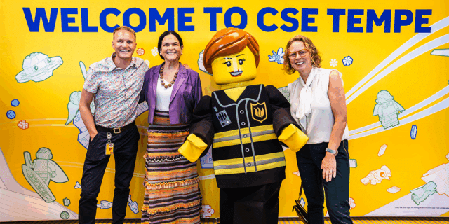 Three people stand around a life-size, humanoid LEGO in front of a yellow wall with hte message "Welcome to CSE Tempe."