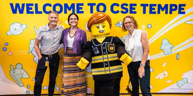 Three people stand around a life-size, humanoid LEGO in front of a yellow wall with hte message "Welcome to CSE Tempe."