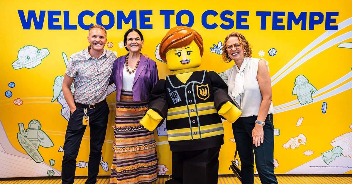 Three people stand around a life-size, humanoid LEGO in front of a yellow wall with hte message "Welcome to CSE Tempe."