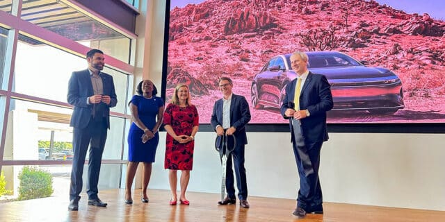 Three men and two women stand on a brown floor. Behind them, an image of a Lucid car in a desert is on a screen.
