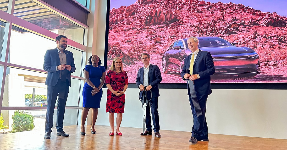 Three men and two women stand on a brown floor. Behind them, an image of a Lucid car in a desert is on a screen.