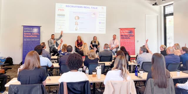 About 20 people sit at tables facing a panel of four speakers. A powerpoint slide is projected onto the wall behind the speakers.