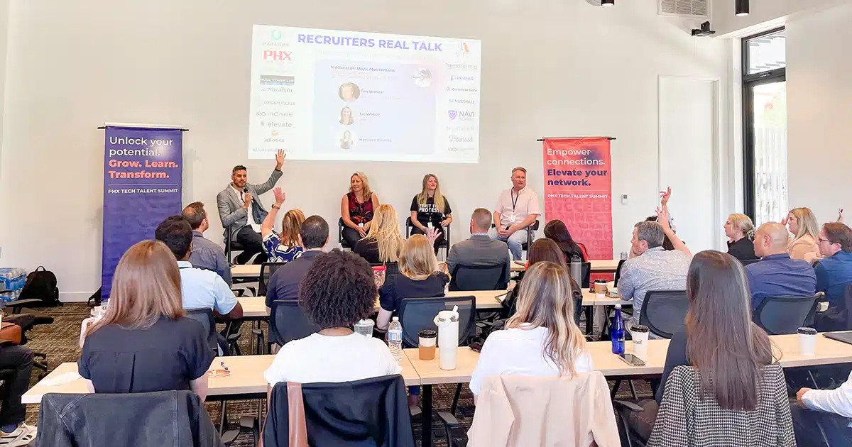 About 20 people sit at tables facing a panel of four speakers. A powerpoint slide is projected onto the wall behind the speakers.
