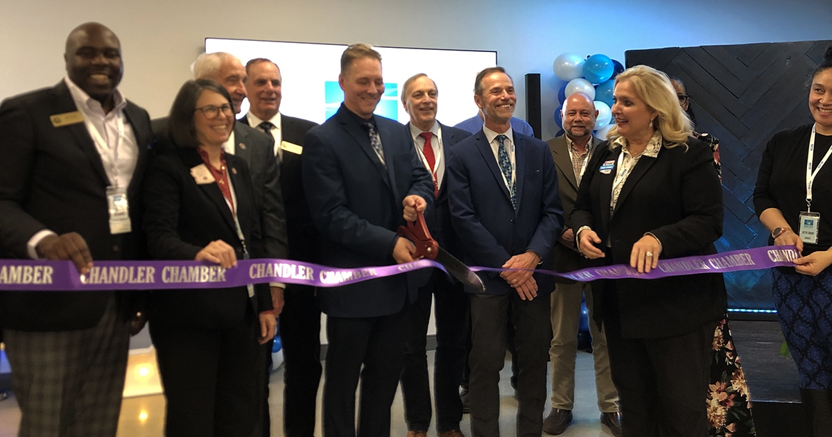 A group of 12 people bunch behind a purple ribbon with an oversized pair of red scissors to celebrate the grand opening of Saras Micro Devices.