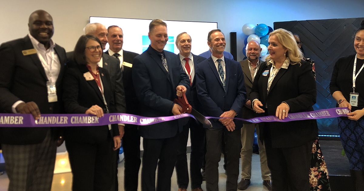 A group of 12 people bunch behind a purple ribbon with an oversized pair of red scissors to celebrate the grand opening of Saras Micro Devices.