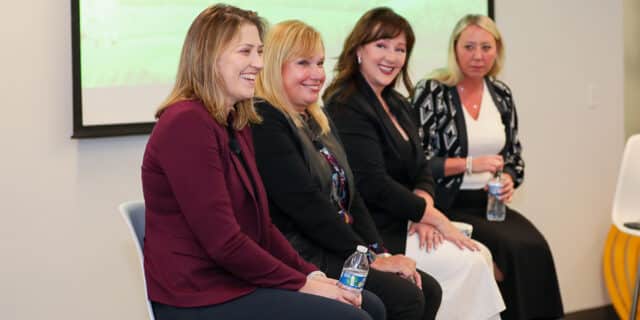 Four women sit in a 45-degree line smiling at an audience off-camera.