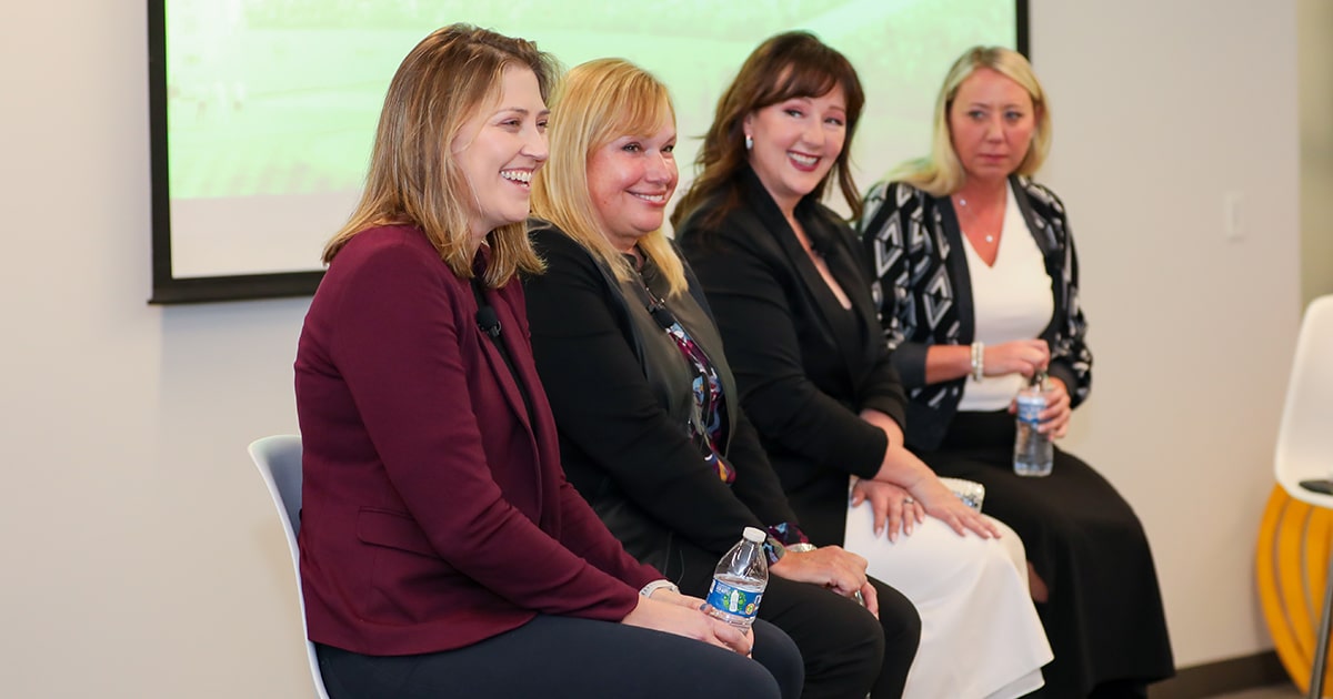 Four women sit in a 45-degree line smiling at an audience off-camera.