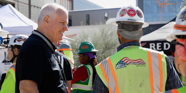 A man facing 90 degrees talks to a man in a construction vest whose back faces the camera.