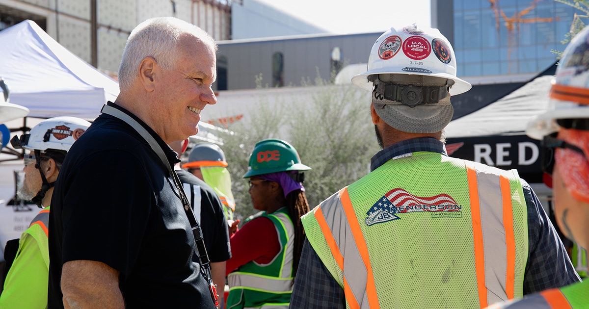 A man facing 90 degrees talks to a man in a construction vest whose back faces the camera.