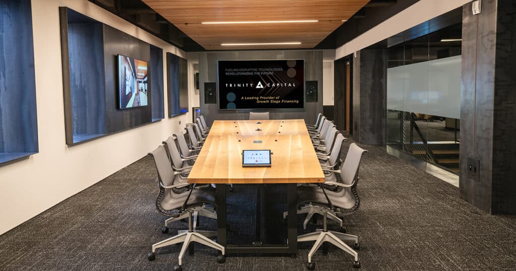 A long conference room table with several chairs around it. At the head is a tv that reads Trinity Capital.