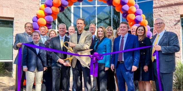 A group of people in professional business attire pose with a giant pair of scissors, prepared to cut a large purple ribbon. A purple-and-orange balloon arch is in the background.