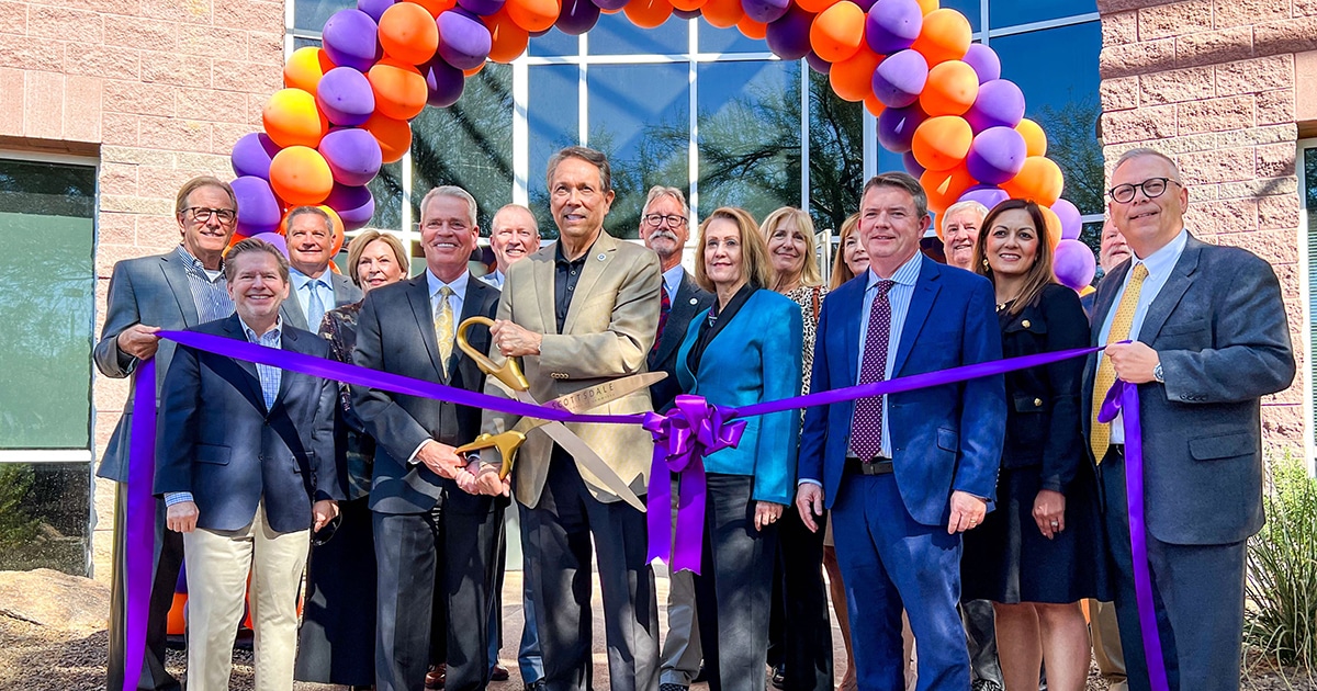 A group of people in professional business attire pose with a giant pair of scissors, prepared to cut a large purple ribbon. A purple-and-orange balloon arch is in the background.