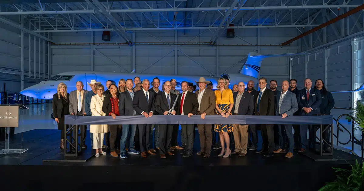 A gaggle of people stand behind a thick dark ribbon, with the person in the middle holding giant scissors. Behind them is an airplane.